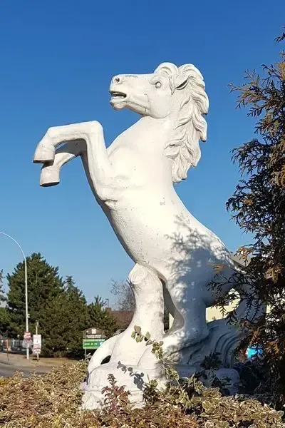 Eine weiße Pferdestatue steht auf zwei Beinen mit einem erhobenen Vorderbein. Sie ist von grünen Büschen und Bäumen umgeben. Der Himmel ist klar und blau.