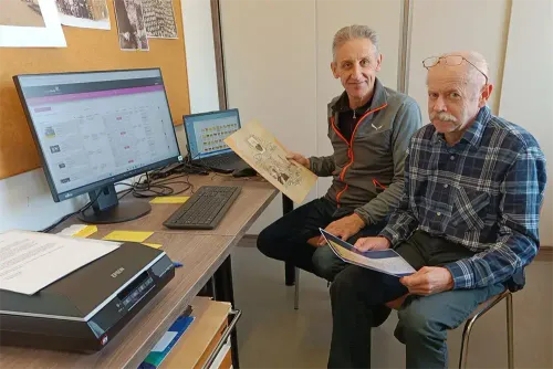 Zwei ältere Männer sitzen an einem Schreibtisch, einer hält ein Papier in der Hand und der andere liest ein Buch. Auf dem Schreibtisch befinden sich ein Monitor, eine Tastatur, eine Maus und ein Drucker. Ein Laptop steht ebenfalls auf dem Schreibtisch. An der Wand hinter ihnen hängen einige Poster.