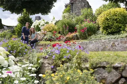 Zwei Frauen stehen in einem Garten voller bunter Blumen, üppiger Grünpflanzen und einer Steinmauer. Sie lächeln und posieren für ein Foto.
