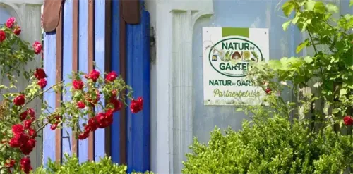 Ein Schild für die Partnerschaft 'Natur-Garten' hängt an einer Wand, umgeben von roten Rosen und grünem Laub.
