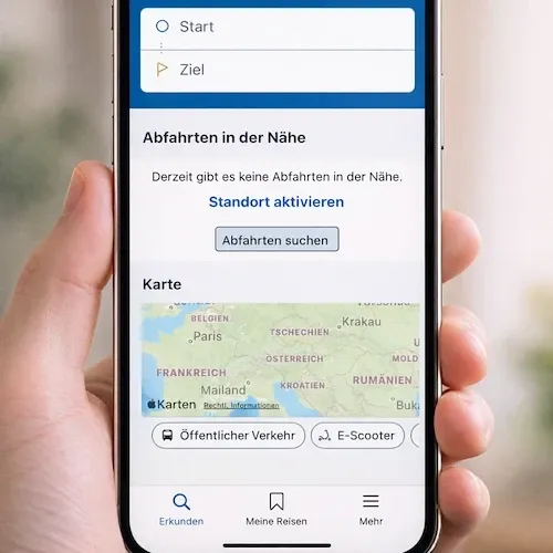 Eine Person hält ein Smartphone mit einer Karte verschiedener Orte. Es gibt Buttons für 'Öffentlicher Verkehr' und 'E-Scooter' sowie Optionen für 'Entdecken', 'Meine Reisen' und 'Mehr'. Die Karte zeigt Regionen wie Belgien, Frankreich und Deutschland.