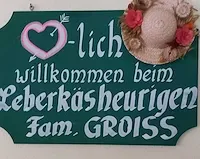 Ein grünes Schild mit einem rosa Herz und Pfeil, einem Hut mit Blumen und den Worten 'Herzlich willkommen beim Leberkasseurigen Fam. GROISS' in weiß.