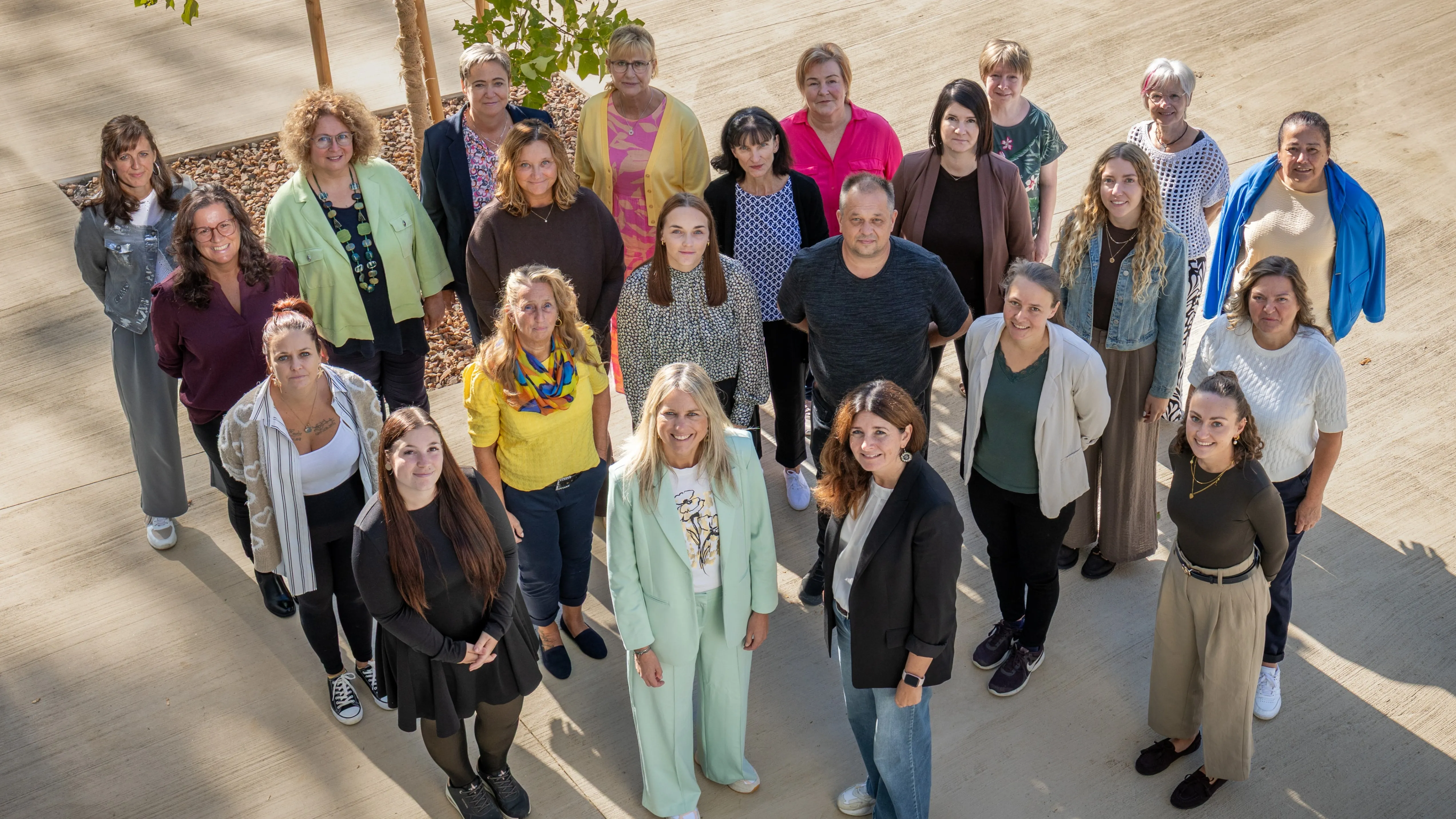 Eine Gruppe von Erwachsenen, sowohl Männer als auch Frauen, stehen im Kreis auf einer Betonfläche, möglicherweise für ein Gruppenfoto.