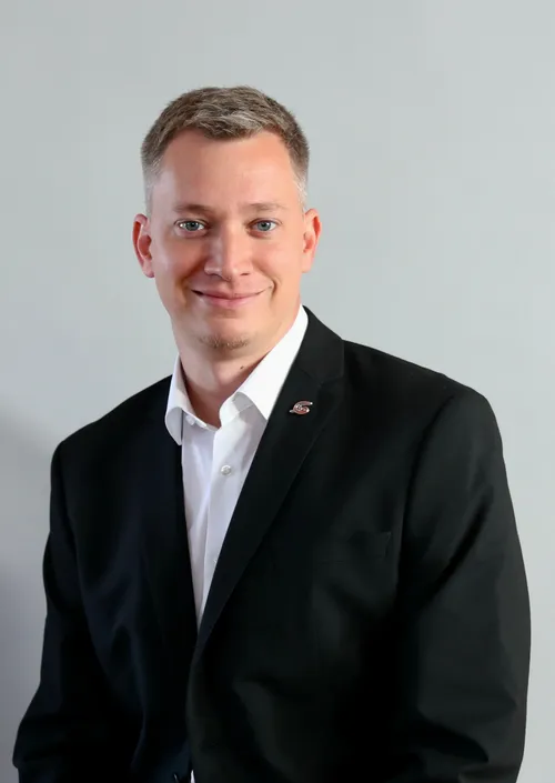 Ein Mann in einem schwarzen Anzug und einem weißen Hemd mit einer Plakette auf der linken Brust lächelt in die Kamera.