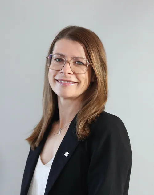 Eine Frau mit braunen Haaren, die eine Brille und einen schwarzen Blazer mit einem weißen Unterhemd trägt. Sie lächelt und schaut direkt in die Kamera.