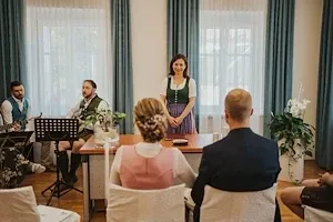 Eine Gruppe von Menschen sitzt um einen Tisch in einem Raum. Eine Frau steht vor dem Tisch, und ein Mann spielt Gitarre. Topfpflanzen stehen auf beiden Seiten des Raumes.