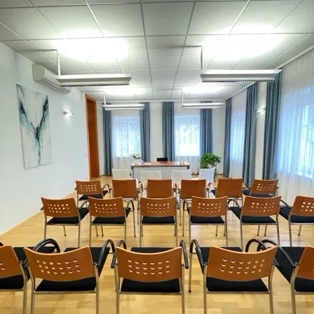 Ein Konferenzraum mit Reihen von Stühlen, einem Tisch mit einem Monitor und einem Gemälde an der Wand. Der Raum hat eine weiße Wand, einen Holzboden und Deckenleuchten.