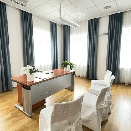 Ein Raum mit einem Schreibtisch und vier Stühlen. Der Schreibtisch hat eine weiße Blumenvase und eine Pflanze darauf. Der Raum hat Holzböden, weiße Wände und blaue Vorhänge.