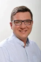 Ein lächelnder Mann mit Brille steht vor einem schlichten weißen Hintergrund und trägt ein hellblaues Hemd mit Knöpfen.