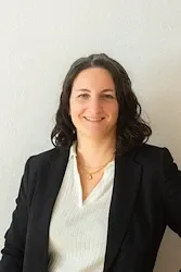 Eine Frau mit kurzen dunklen Haaren, lächelnd, steht vor einer weißen Wand. Sie trägt einen schwarzen Blazer und ein weißes Oberteil.