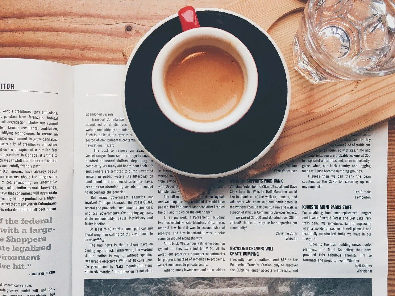 Eine Tasse Kaffee auf einer Untertasse steht auf einem offenen Buch auf einem Holztisch. Ein Glas Wasser und eine Holzwand sind im Hintergrund.