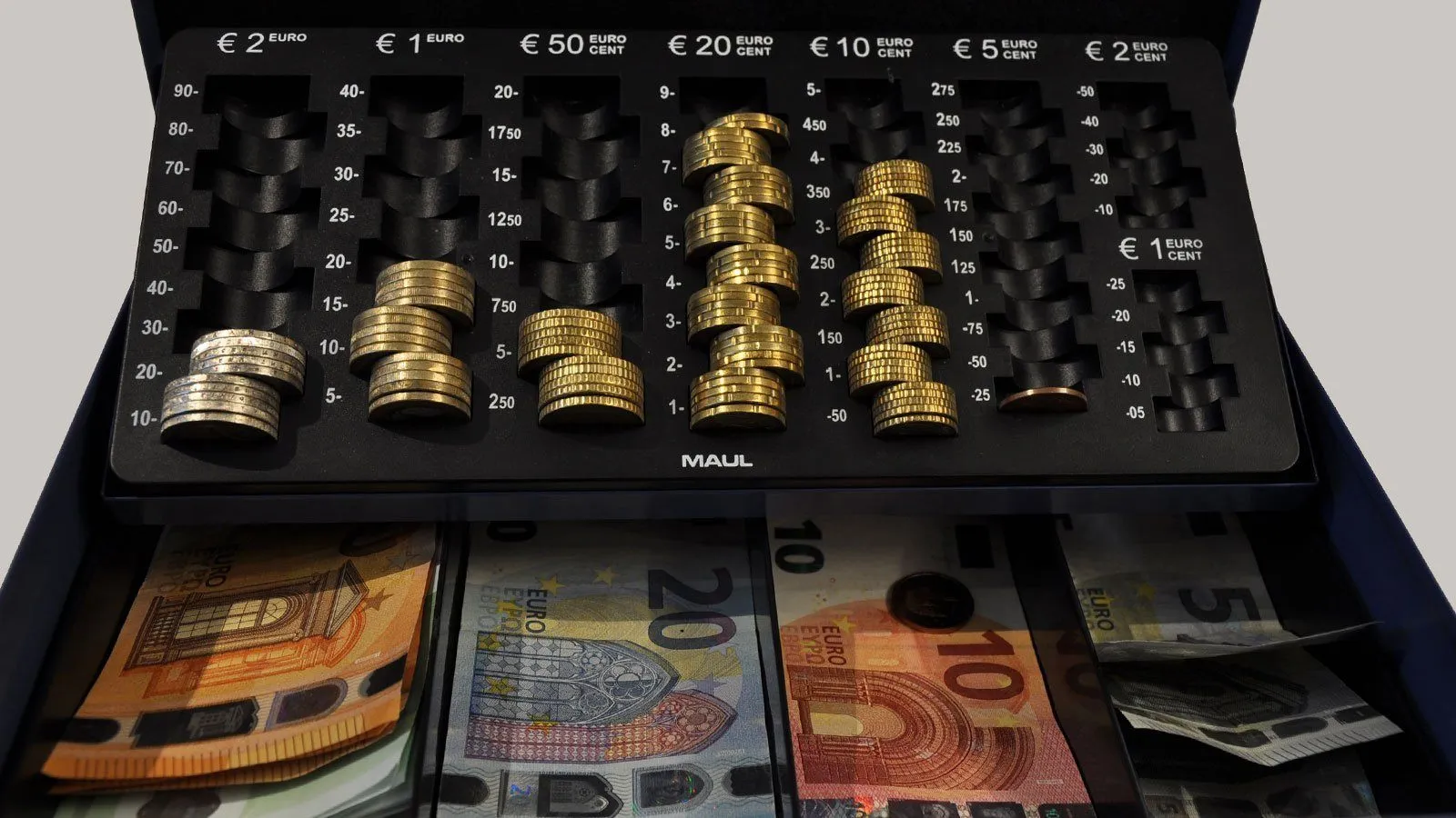 Eine Nahaufnahme einer Münzzählmaschine mit verschiedenen Goldmünzen und Euro-Banknoten. Die Maschine hat Fächer, die mit Zahlen und Münzwerten beschriftet sind.
