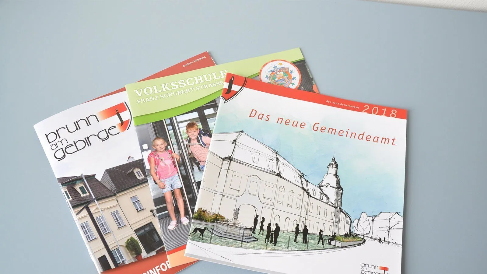 Drei Broschüren sind auf einem blauen Hintergrund zu sehen. Die oberste Broschüre trägt den Titel 'Volksschule Franz Schubert Strasse'. Die mittlere heißt 'Das neue Gemeinde'. Die untere ist teilweise sichtbar.