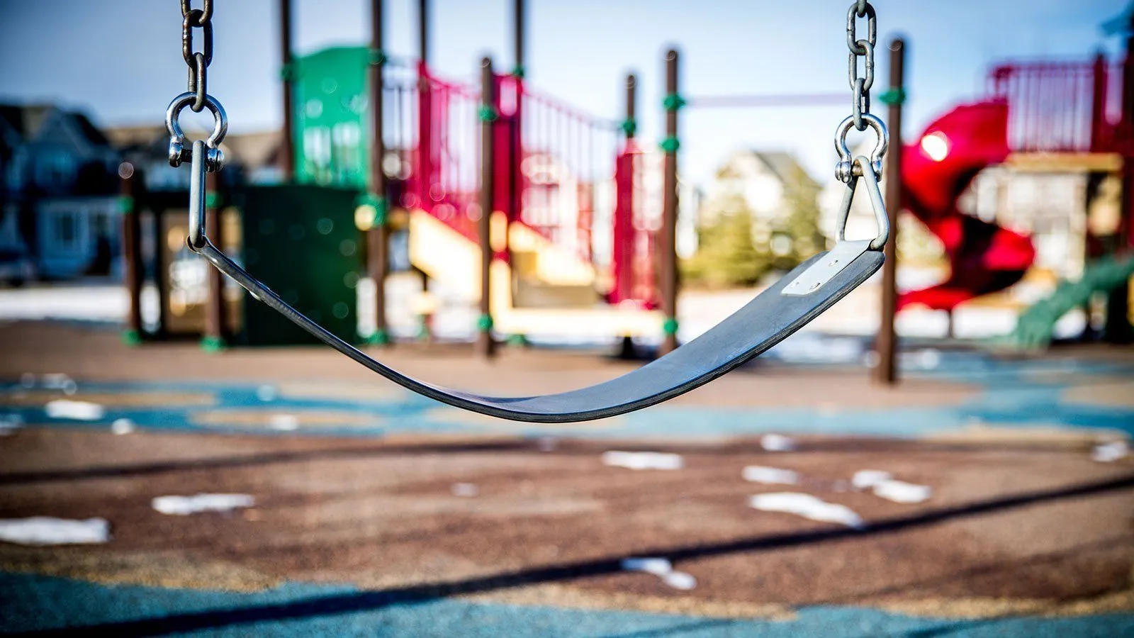 Ein Spielplatz-Szene mit einem Schaukelset im Fokus, bunte Spielstrukturen und eine Rutsche im Hintergrund, unter einem klaren Himmel.