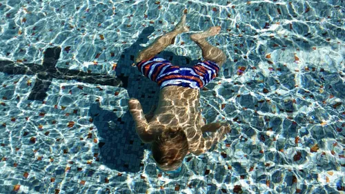 Ein Kind schwimmt kopfüber in einem Schwimmbecken und trägt bunte Badeshorts. Der Poolboden ist gefliest.