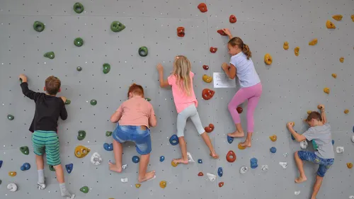 Drei Kinder klettern an einer Wand mit unterschiedlich farbigen Griffen. Ein Kind liegt auf dem Rücken, während die anderen auf den Zehenspitzen stehen.