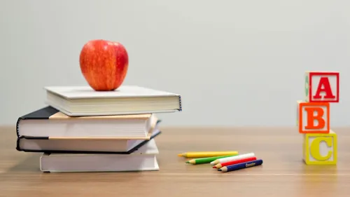 Ein roter Apfel sitzt auf einem Stapel Bücher auf einem Tisch. Verschiedene farbige Stifte liegen verstreut auf dem Tisch.