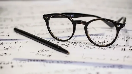 Schwarze Brille liegt auf einem Musikblatt mit einem Stift daneben. Das Blatt hat Noten und Pausen.