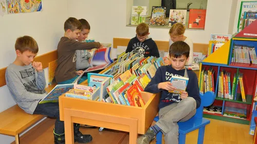 Mehrere Kinder sind in einer Bibliothek. Sie stöbern in Büchern. Die Kinder sitzen auf einem blauen Stuhl und auf einem Regal.