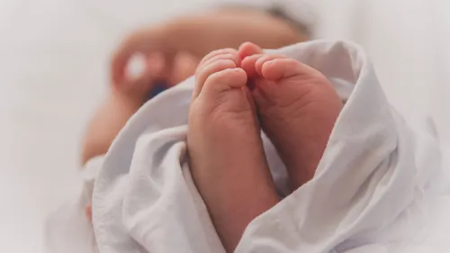 Nahaufnahme der Füße eines Neugeborenen, eingewickelt in eine weiße Decke. Die Hände des Babys sind leicht gekrümmt.