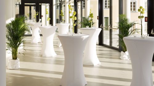 Mehrere weiße zylindrische Tische mit Blumenarrangements sind in einem modernen, offenen Raum mit Glastüren aufgereiht. Die Tische haben Glasplatten und der Raum hat weiße Wände und Böden.