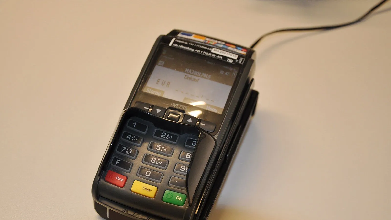 Ein schwarzer Zahlungsterminal steht auf einem Tisch, dessen Bildschirm 'EUR' anzeigt. Das Gerät hat eine Tastatur mit Zahlentasten und farbigen Tasten wie rot, gelb und grün.