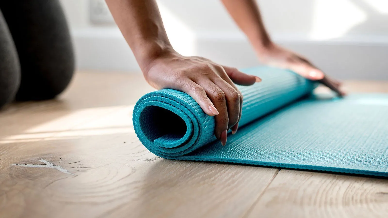 Eine Hand entrollt eine türkisfarbene Yogamatte auf einem Holzboden und zeigt deren strukturierte Oberfläche. Die Matte ist ordentlich aufgerollt.