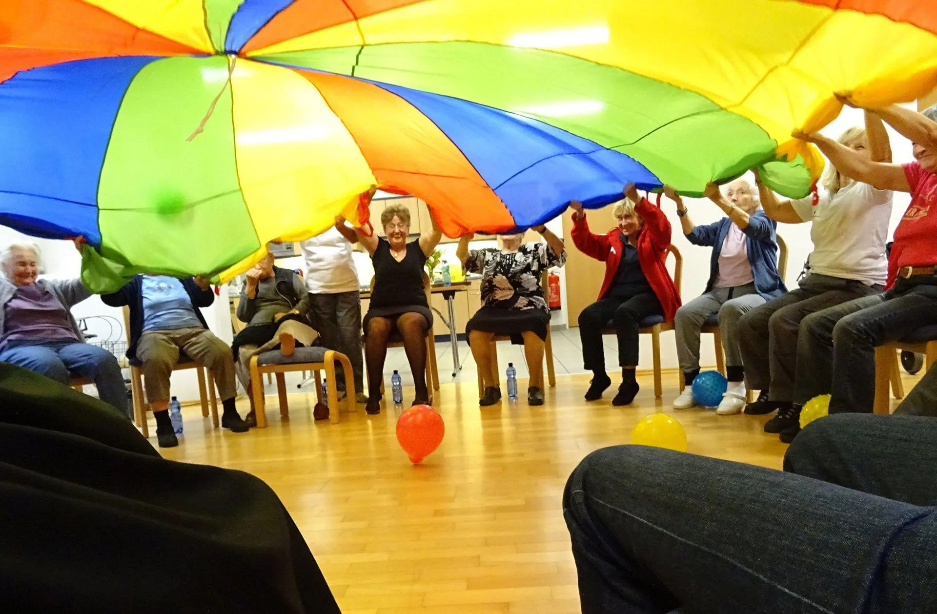 Eine Gruppe älterer Erwachsener sitzt auf Stühlen und hält einen großen bunten Fallschirm hoch. Ein roter Ballon schwebt in der Nähe, während andere auf dem Boden sind.