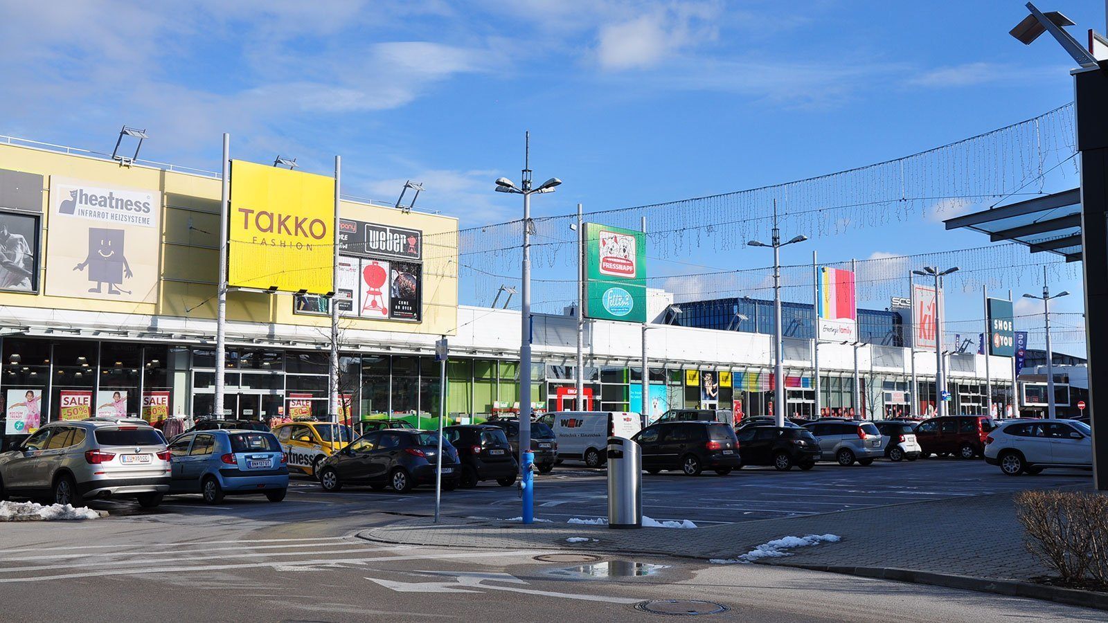 Ein Parkplatz mit vielen geparkten Autos vor verschiedenen Geschäften, darunter Takko Fashion, Weber und Fit&Fresh. Straßenlaternen und ein Mülleimer sind sichtbar.