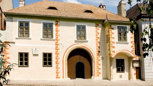 Ein weißes Gebäude mit einem braunen Dach, einem hölzernen Bogentor und drei Fenstern. Das Bogentor ist aus Holz und hat ein aufwendiges Design.