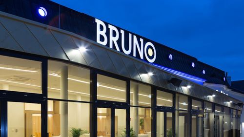 Die Vorderseite eines Gebäudes mit einem großen weißen Schild mit der Aufschrift Bruno. Es hat Glastüren und Fenster, die von Lichtern beleuchtet sind. Das Gebäude hat ein modernes Design.