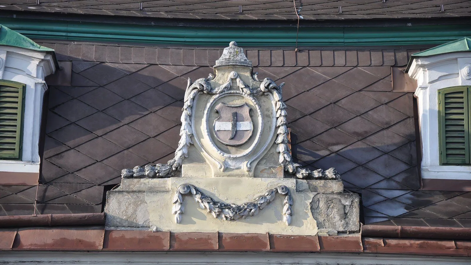 Ein kunstvolles Wappen schmückt die Spitze eines Gebäudes. Es zeigt einen Schild mit einem Kreuz und dekorativen Verzierungen.