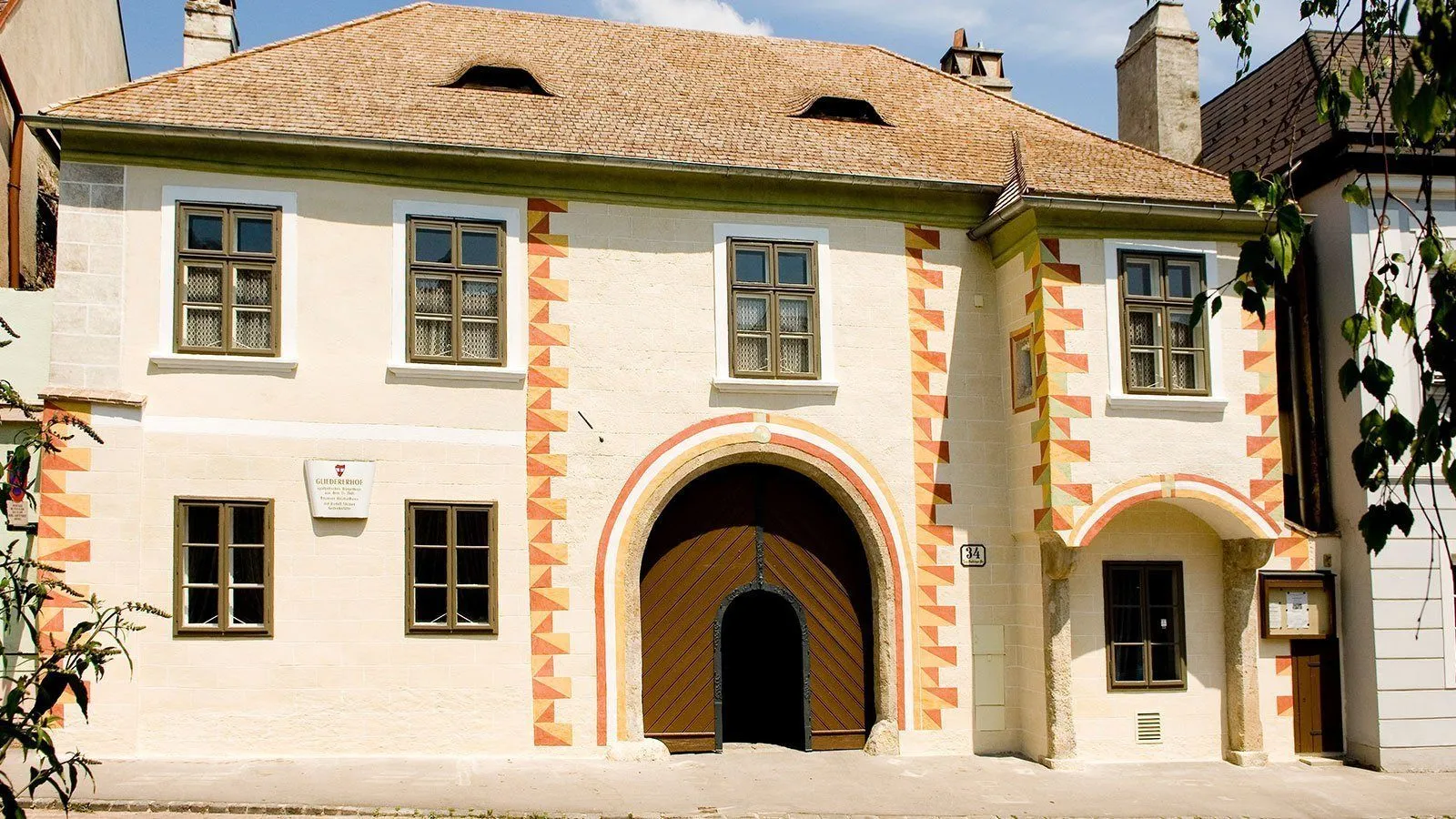 Ein altes Gebäude mit einem braunen Holztor, einem gewölbten Eingang und gemusterten Designs an den Wänden. Es gibt drei Fenster und ein kleines Schild mit Schrift auf der linken Seite.