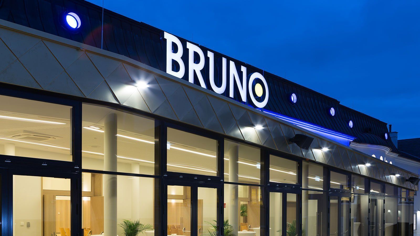 Das Äußere eines modernen Gebäudes, Bruno, mit Glaswänden, nachts beleuchtet und blauem Licht auf dem Dach.