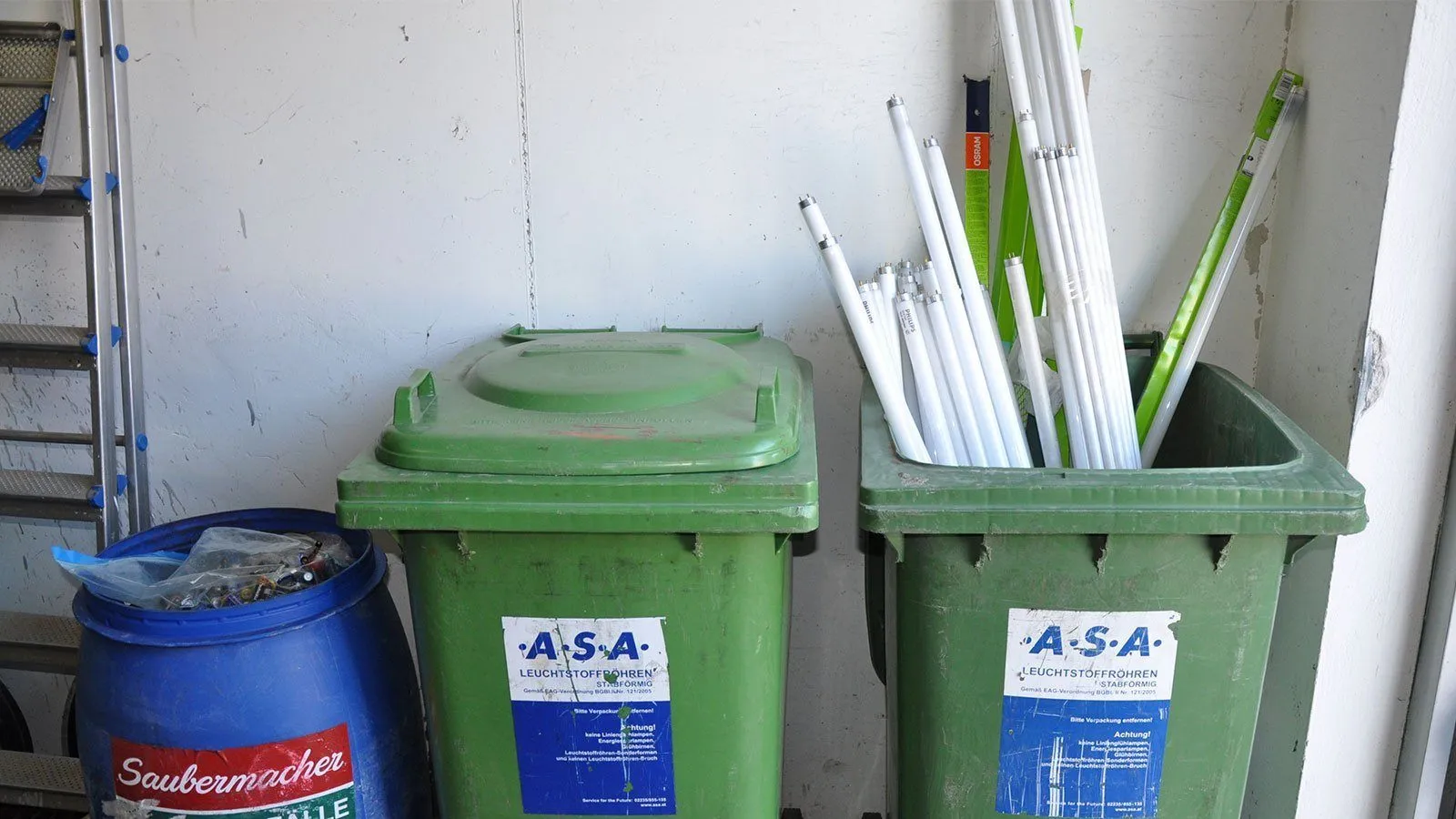 Zwei grüne Behälter sind gegen eine Wand gestapelt. Einer hat ein Etikett mit der Aufschrift ASA und der andere ist mit Leuchtstoffröhren gefüllt.