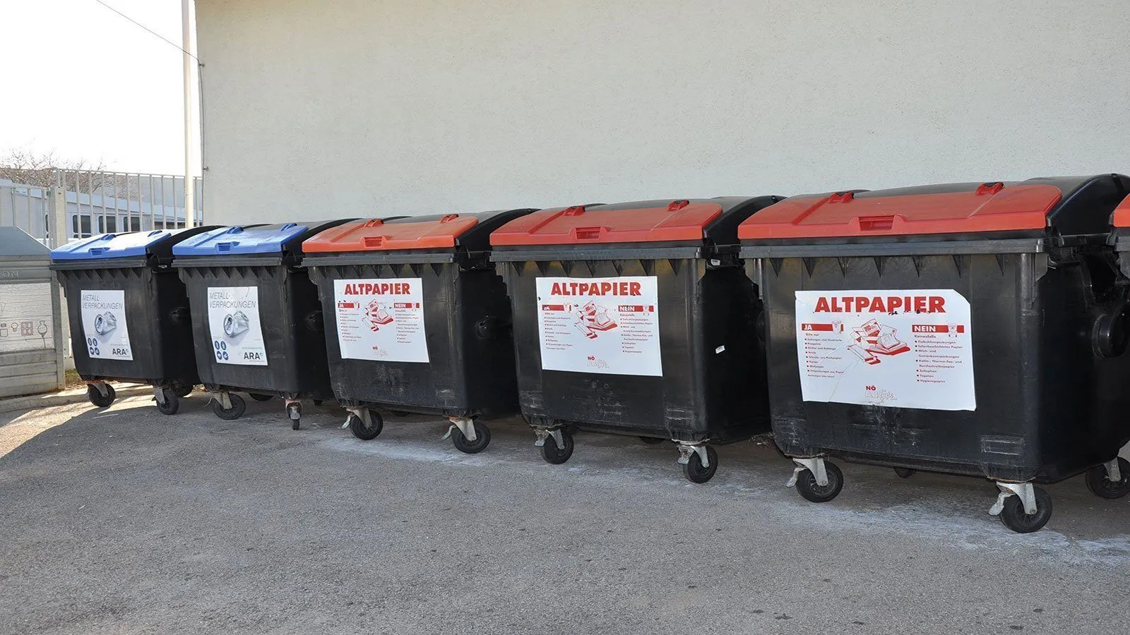 Vier schwarze Recycling-Behälter mit roten Deckeln, beschriftet mit 'Altpapier', sind auf einer Betonfläche aufgereiht. Jeder Behälter hat einen Aufkleber mit Anweisungen.