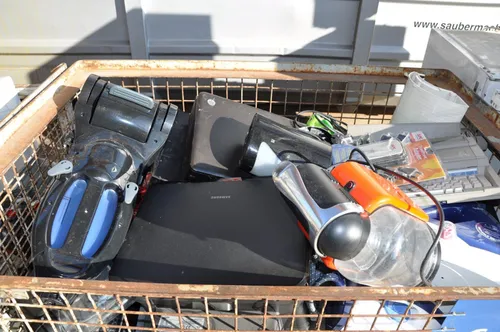 Ein Einkaufswagen gefüllt mit verschiedenen Elektronikgeräten, darunter ein Samsung-Laptop, ein schwarzer Staubsauger und ein schwarzes und silbernes Gerät.