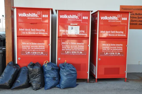 Drei rote Recycling-Behälter sind mit blauen Säcken, die Kleidung enthalten, davor aufgereiht. Die Behälter haben weiße Etiketten mit dem Volkshilfe.Box-Logo und Text auf Deutsch.