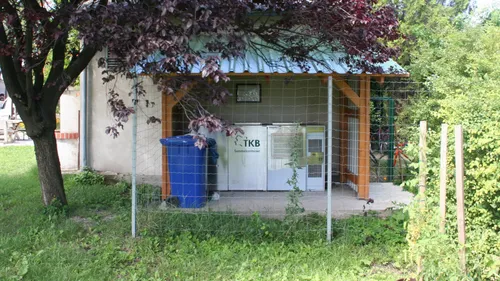 Ein überdachter Außenbereich mit einem blauen Mülleimer, zwei weißen Behältern und einem Metallzaun. Über den Behältern befindet sich ein Schild mit den Buchstaben 'TKB'. Ein Baum mit violetten Blättern bedeckt teilweise den Eingang.