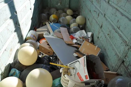 Ein Müllcontainer voller Abfall, einschließlich zerbrochener Lampen, Kisten, Plastiktüten und einem weißen Korb. Der Müllcontainer hat eine grüne Wand.