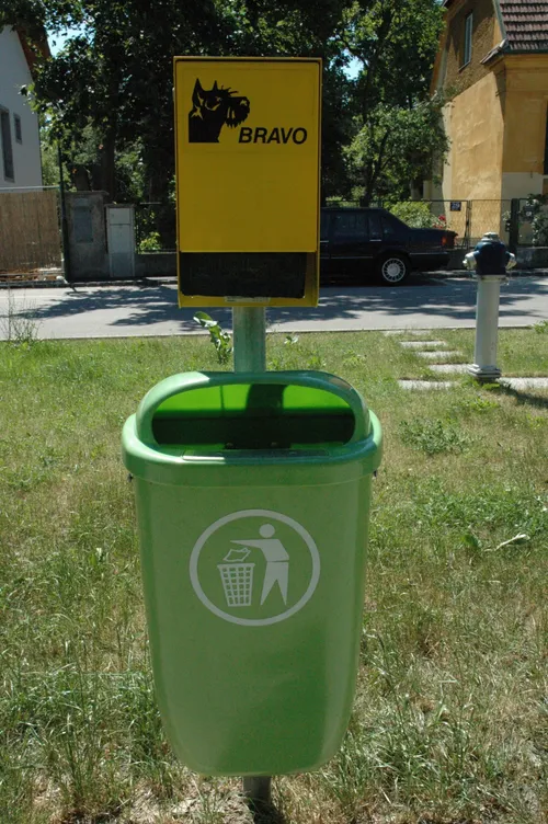 Ein grüner Mülleimer mit einem Schild, das BRAVO steht auf einem Rasen. Dahinter ist ein Feuerhydrant zu sehen, und ein schwarzes Auto ist in der Ferne geparkt.