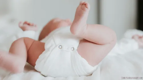 Ein Baby liegt auf einem Bett und trägt einen weißen Stoff mit Knöpfen. Der Fuß des Babys ist angehoben und die Hand leicht gekrümmt.
