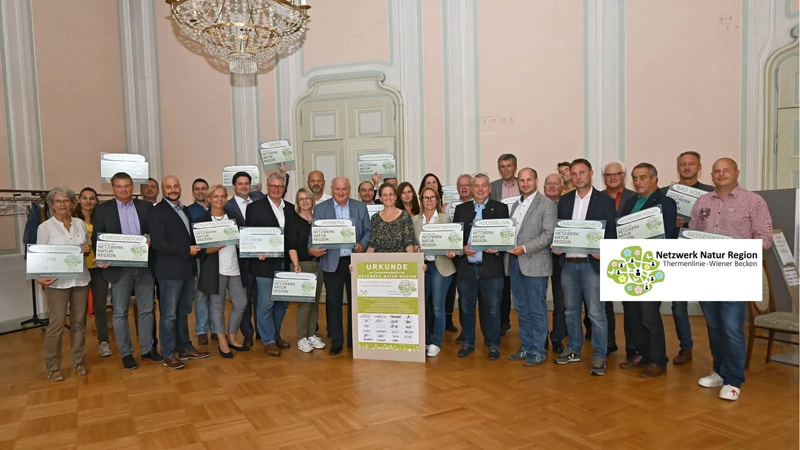 Eine Gruppe von Menschen in Anzügen und Turnschuhen halten Urkunden vor einem Plakat. Sie stehen in einem Raum mit Holzböden, einer Kronleuchter und einer weißen Wand.
