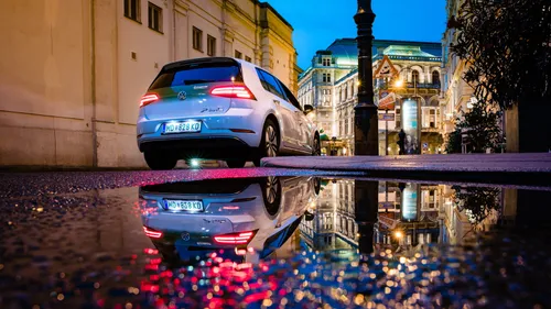 Ein weißer Volkswagen in der Nacht in der Stadt geparkt, sein Spiegelbild in einer Pfütze auf der Straße sichtbar. Die Rücklichter des Autos sind eingeschaltet, und die Umgebung wird von Straßenlaternen und Gebäuden beleuchtet.