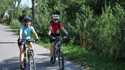 Zwei Personen mit Helmen und Sonnenbrillen fahren auf einem asphaltierten Weg, umgeben von Grün. Die Person vorne trägt ein rot-schwarzes Trikot.