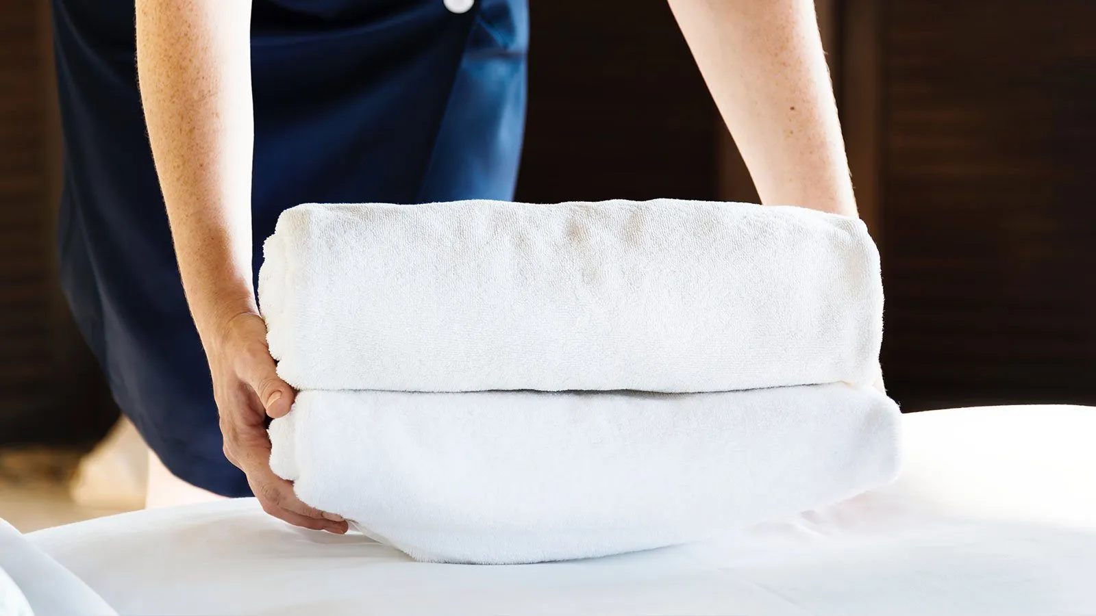 Eine Person in blauer Uniform hält zwei weiße Handtücher auf einer weißen Oberfläche, wahrscheinlich in einem Hotel.