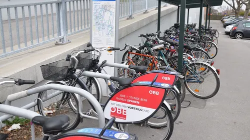 Eine Reihe von Fahrrädern, die an einer Station geparkt sind, mit einer Karte dahinter. Die Fahrräder haben Körbe und Karten mit der Aufschrift OBB.