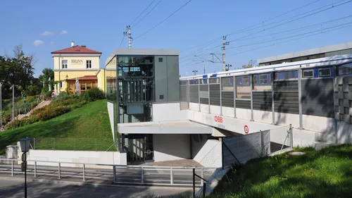 Eingang zu einem Tunnel mit einem Zug im Hintergrund. Der Eingang hat eine Glaswand, Metallgeländer und einen abschüssigen Weg. Es gibt ein Schild im Tunnel mit der Aufschrift 'OBB INFRA'.