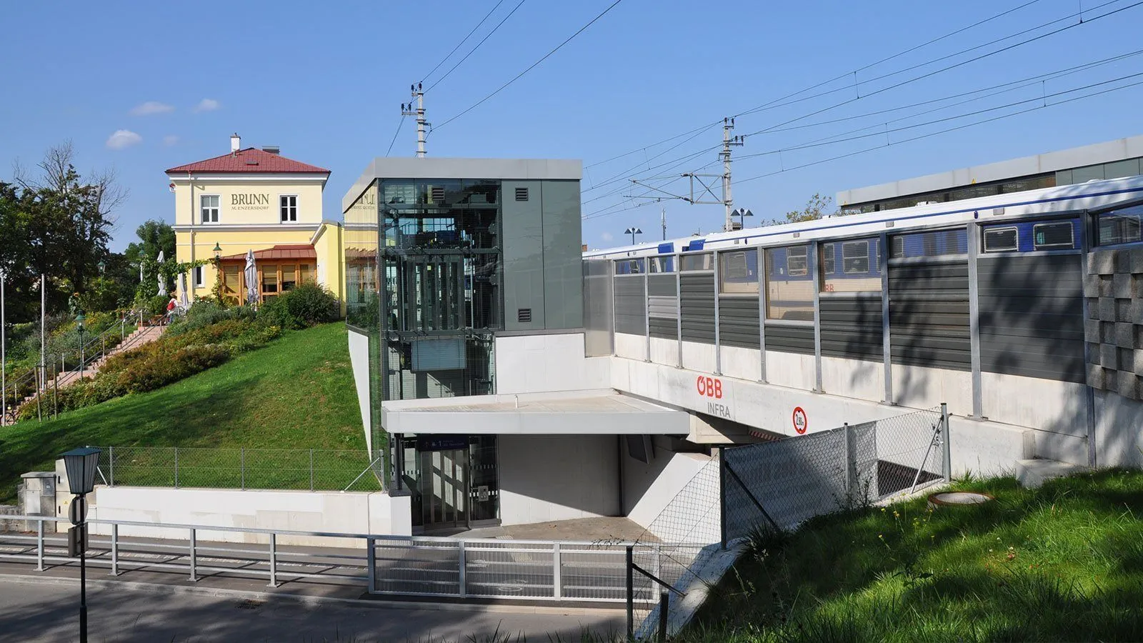 Eine hochgelegene Zugstation mit einem gläsernen Aufzug, der sie mit dem Bodenniveau verbindet. Die Station hat einen grasbewachsenen Hügel und ein Gebäude im Hintergrund.