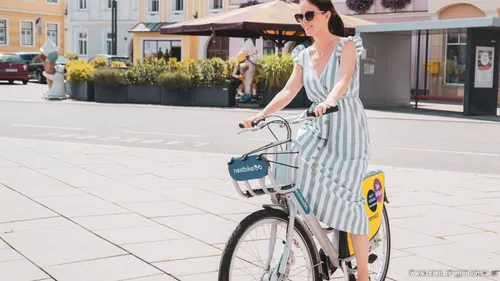 Eine Frau in einem gestreiften Kleid fährt ein Fahrrad mit 'nextbike' vorne. Sie lächelt und steht auf einem Bürgersteig mit einer Statue und Pflanzen im Hintergrund.